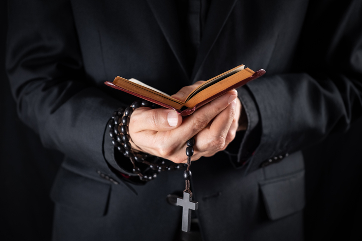 Midsection Of Priest Holding Prayer Beads And Bible While Standing Against Black Background